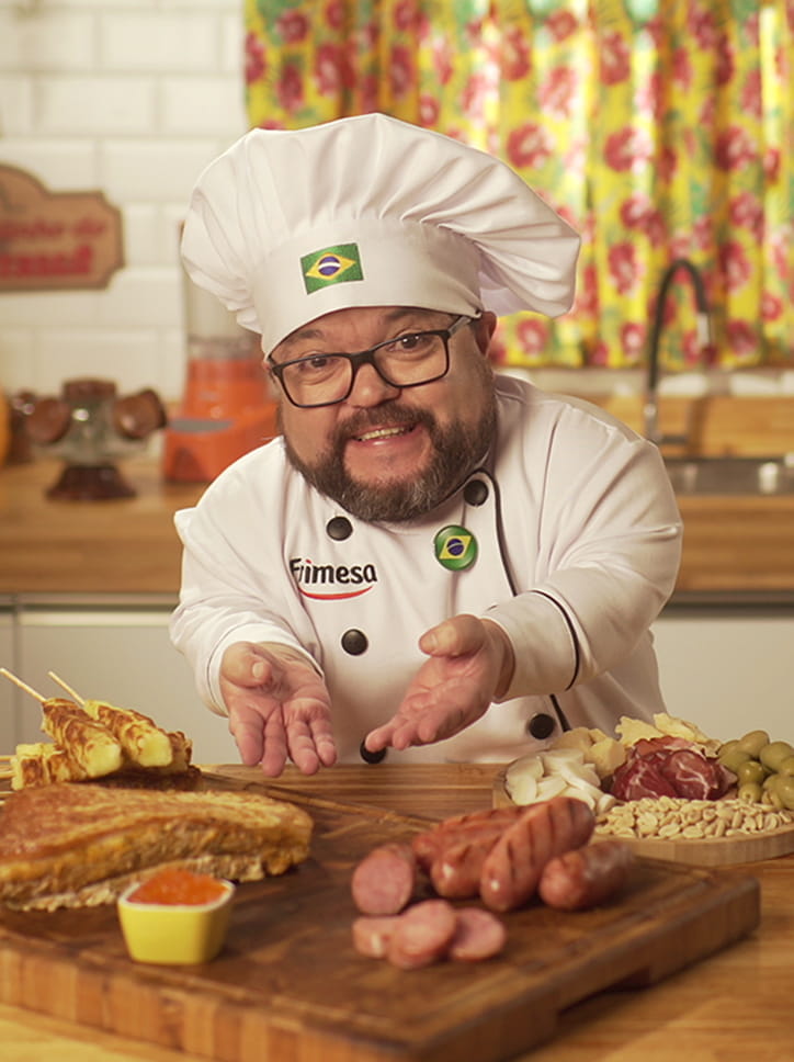 Um chef e humorista em uma peça da campanha da Frimesa, vestido com um dólmã e chapéu com a bandeira do Brasil, apresentando uma tábua de petiscos.