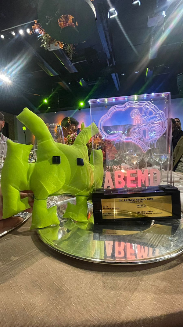 Troféu do 31º Prêmio ABEMD 2025 exibido sobre uma mesa espelhada, ao lado do mascote verde da HouseCricket — o “Grilo”, símbolo da agência. A peça transparente com o logotipo da premiação reflete o prestígio conquistado pela empresa nas categorias de marketing digital e CRM. O ambiente sofisticado do evento reforça o reconhecimento nacional da agência pelo trabalho inovador e criativo.