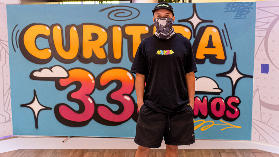 Grafiteiro Diego Prosa posa em frente a um grande painel colorido com a inscrição “Curitiba 333 anos”, criado como parte de uma intervenção artística colaborativa no Shopping Mueller. A ação, idealizada pela HouseCricket, celebra o aniversário da cidade de Curitiba e convida o público a participar da construção coletiva da obra.