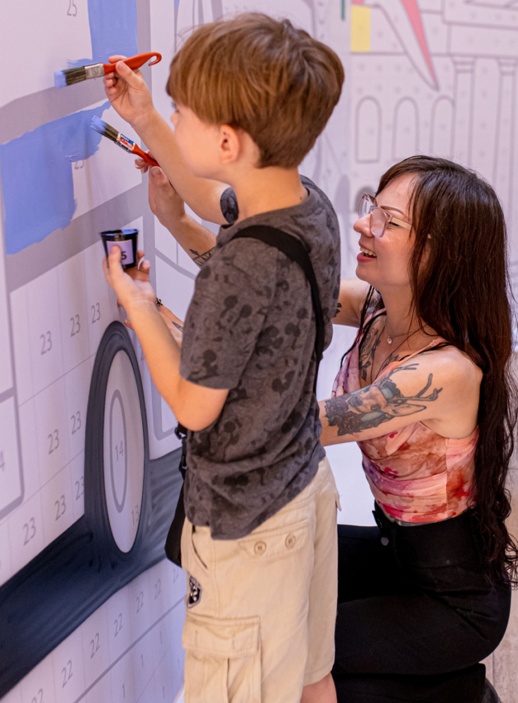 Criança pinta mural colaborativo com apoio de visitante durante ação “Curitiba 333 anos” no Shopping Mueller