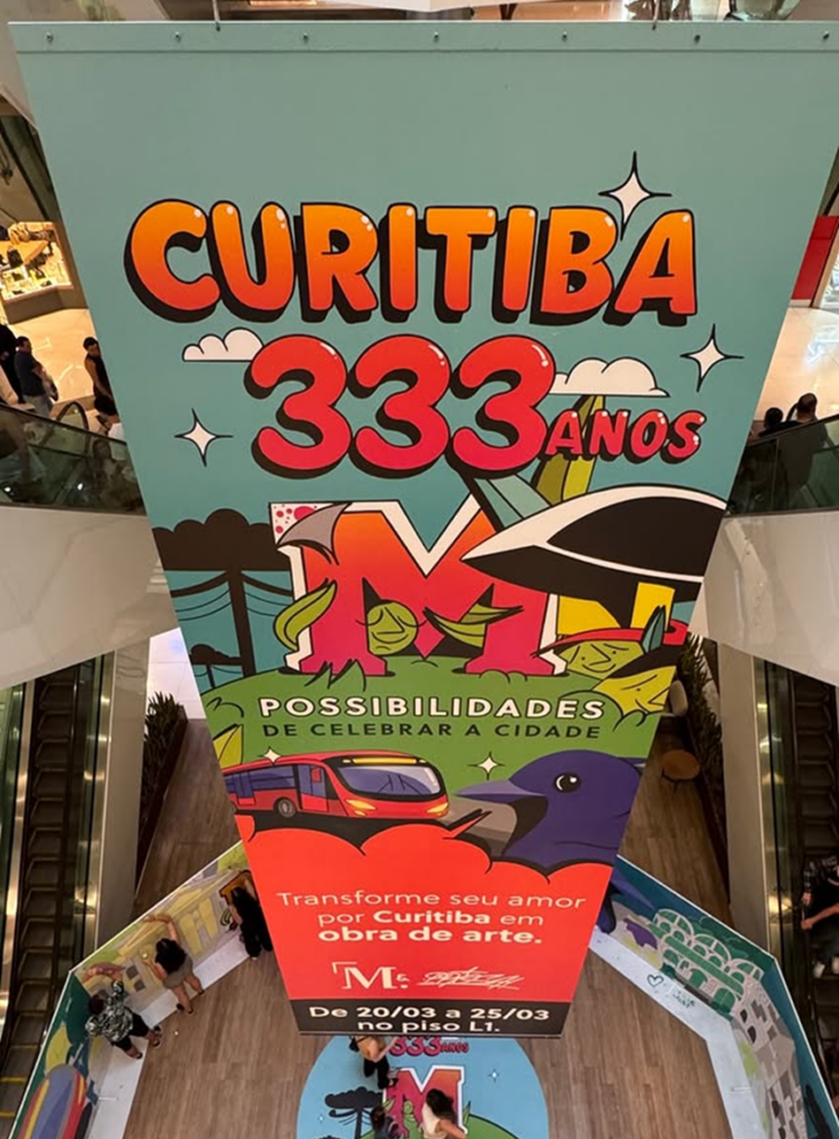 Banner de divulgação da intervenção artística “Curitiba 333 anos” instalado no vão central do Shopping Mueller convida o público a participar da criação do mural colaborativo.