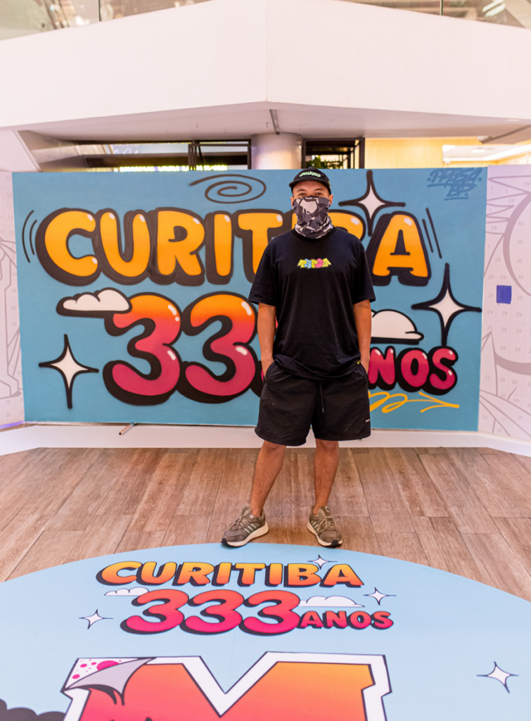 Diego Prosa posa em frente ao painel finalizado “Curitiba 333 anos” no Shopping Mueller, destacando a obra criada de forma colaborativa para celebrar o aniversário da cidade.